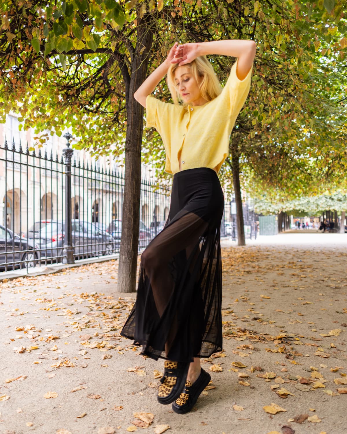 A woman posing outdoors on a tree-lined street, wearing a pale yellow short-sleeve button-up shirt, a high-waisted black midi skirt with sheer side panels, and barefoot loafers with leopard print detailing.