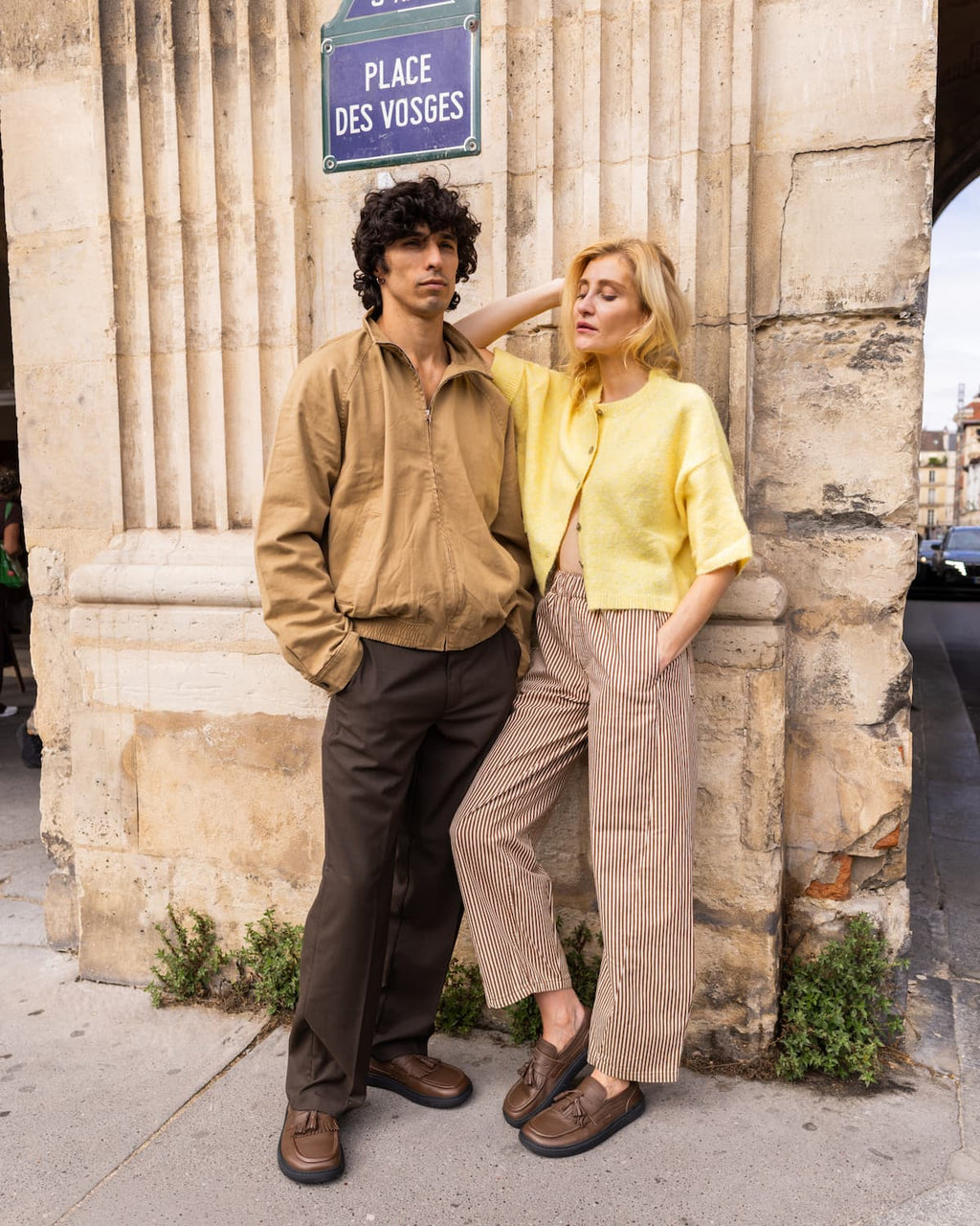 A man and a woman posing outdoors against a stone column, wearing vintage-style casual clothing. The man wears a tan zip-up shirt and dark brown trousers, while the woman wears a yellow cropped cardigan and beige pinstripe trousers.