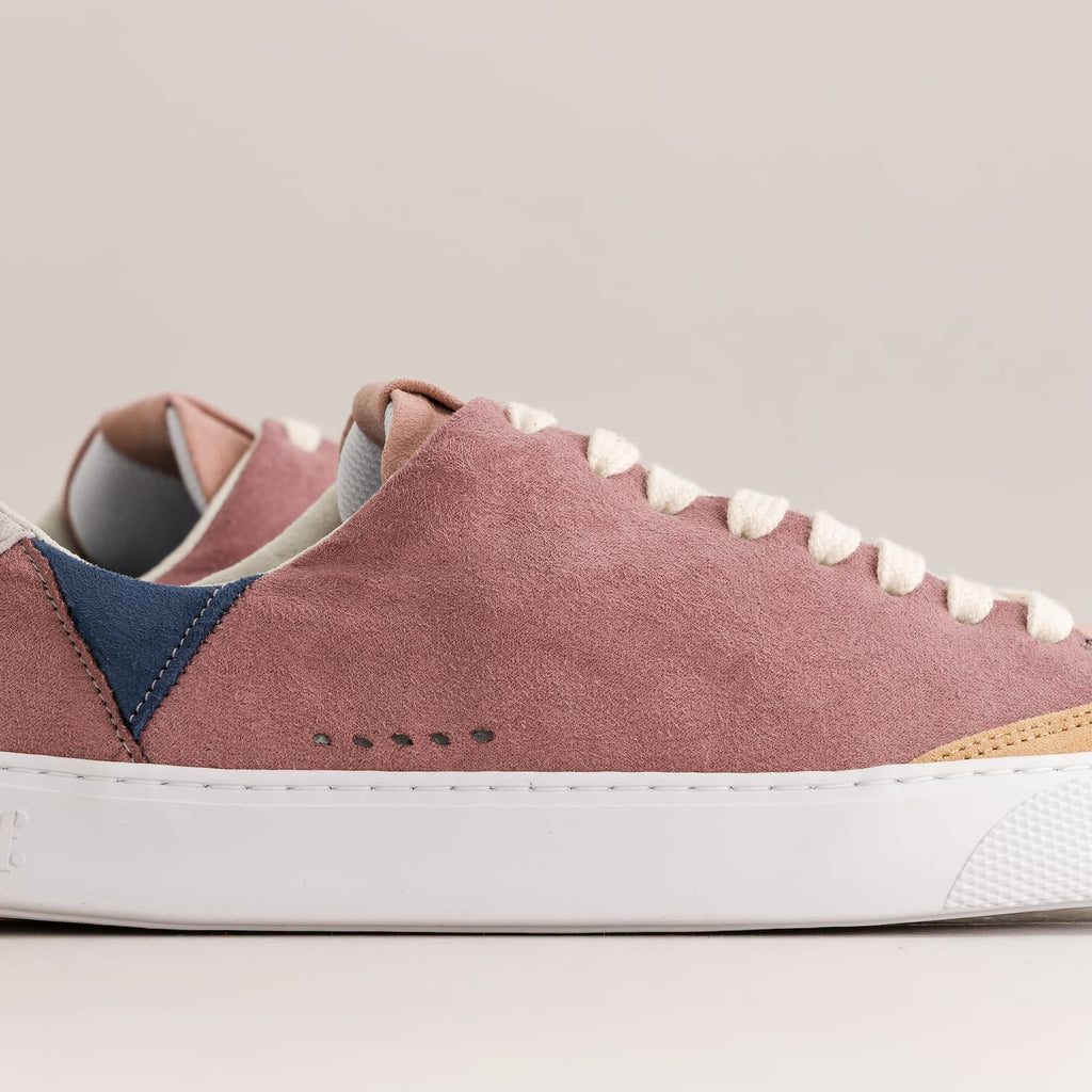 A close-up side view of a barefoot sneaker in dusty rose suede with a navy blue heel panel, white laces, a thick white rubber sole, and a small tan accent near the eyestay.