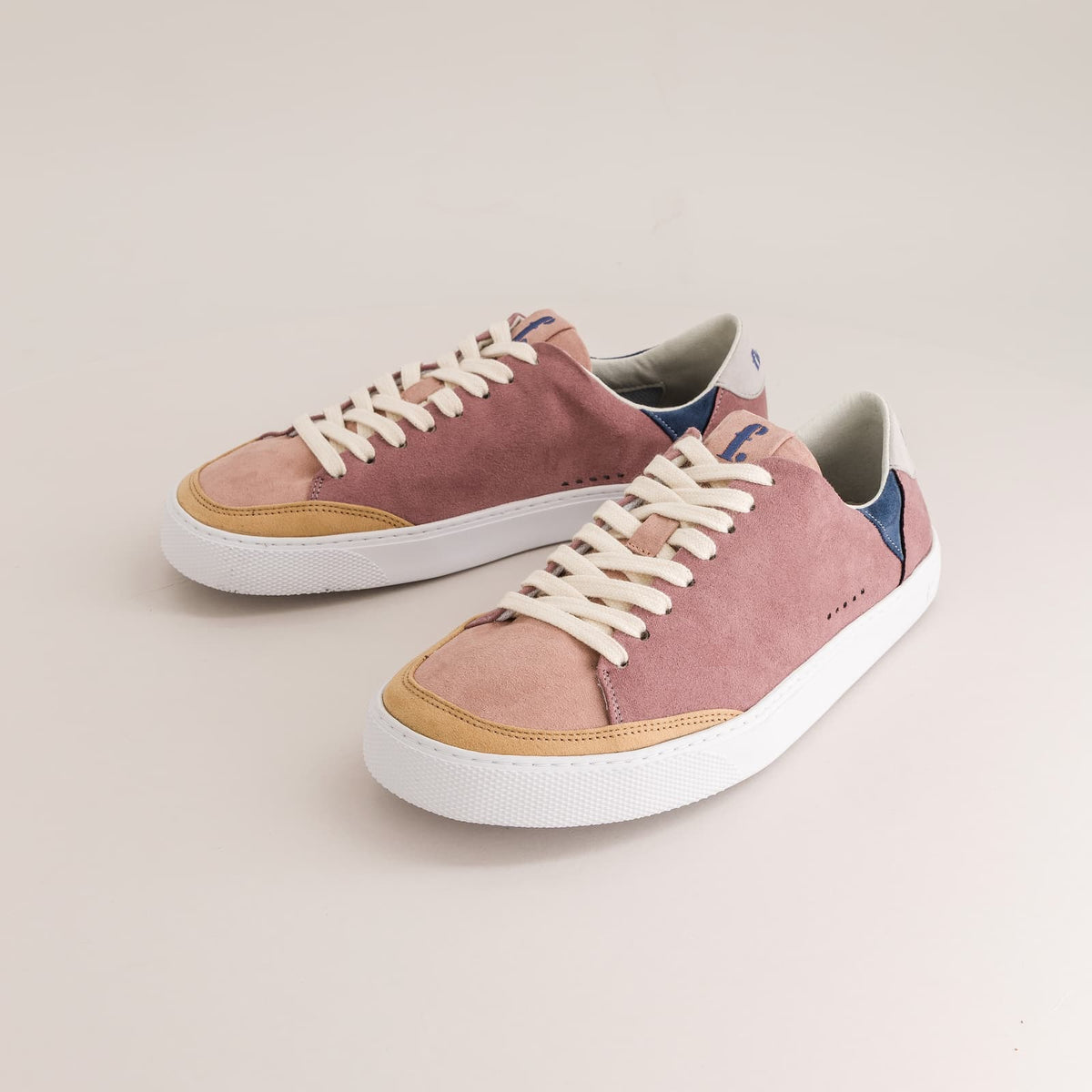 A pair of barefoot sneakers in dusty pink suede with tan leather toe caps, white laces, and white rubber soles, photographed on a light beige background.