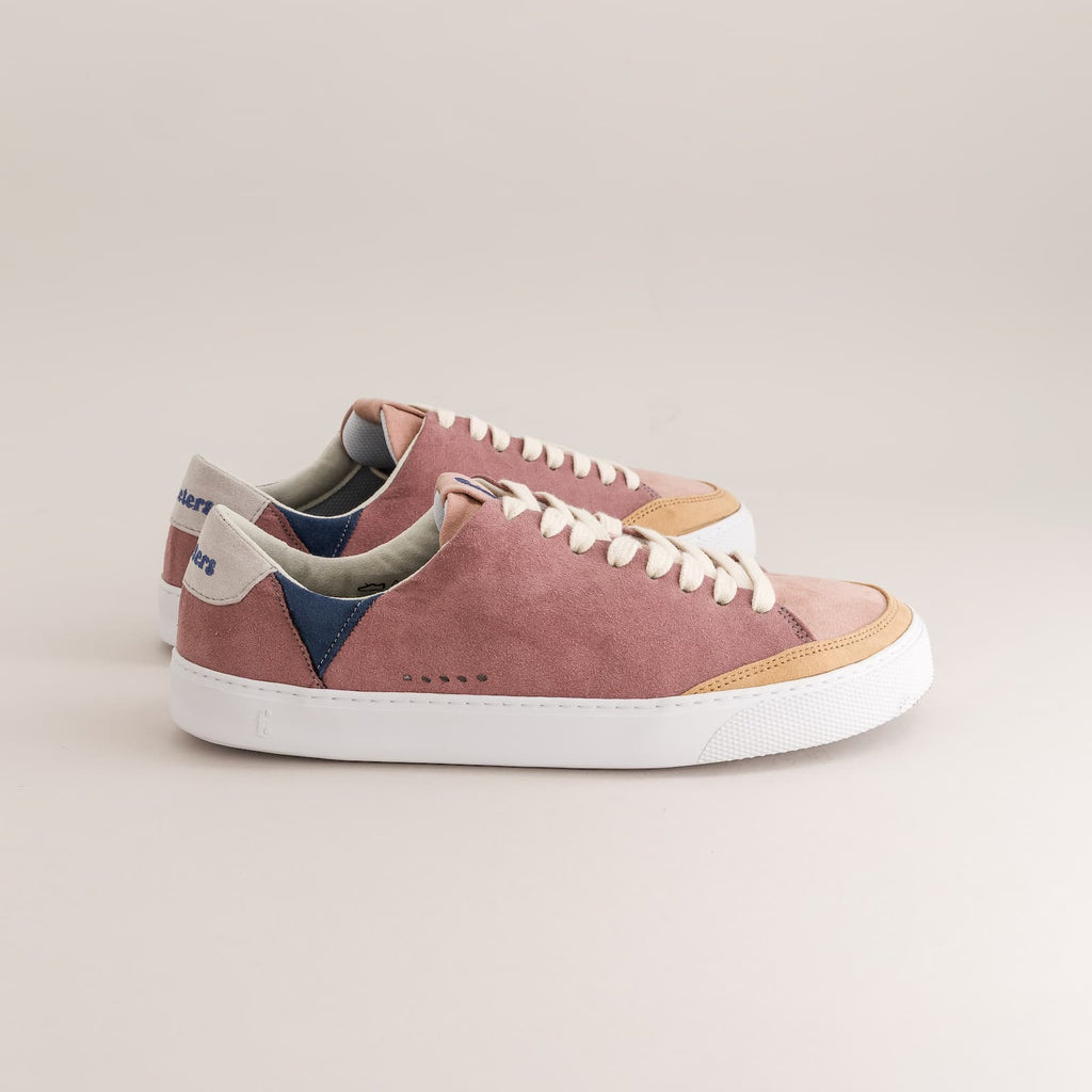 A pair of barefoot sneakers in dusty rose suede with navy blue side panels, white laces, a white rubber sole, and a tan toe cap, photographed on a clean light beige background.