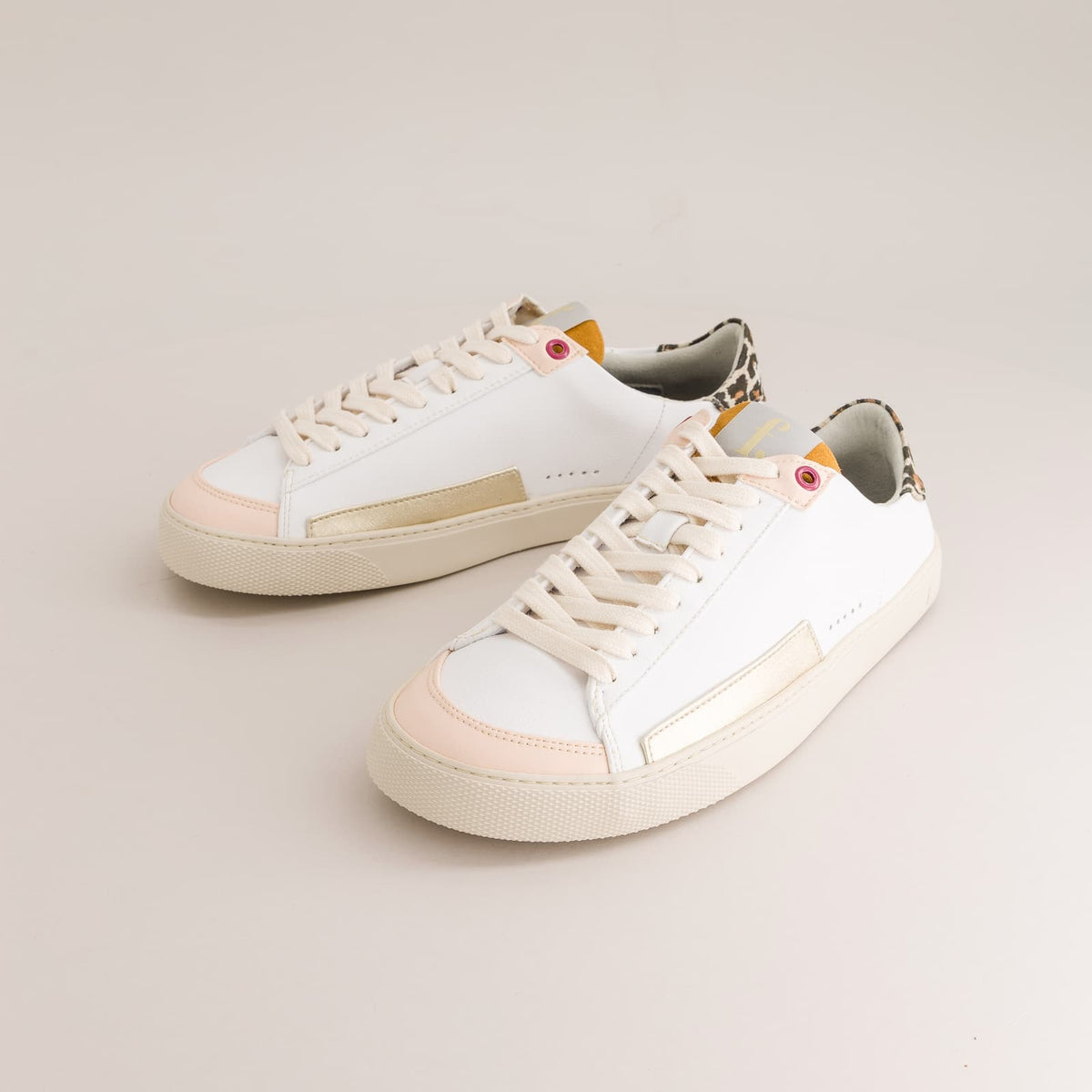 A pair of white leather low-top barefoot sneakers with pale pink toe caps, metallic gold side stripes, and leopard-print heel tabs, photographed on a light beige studio background.