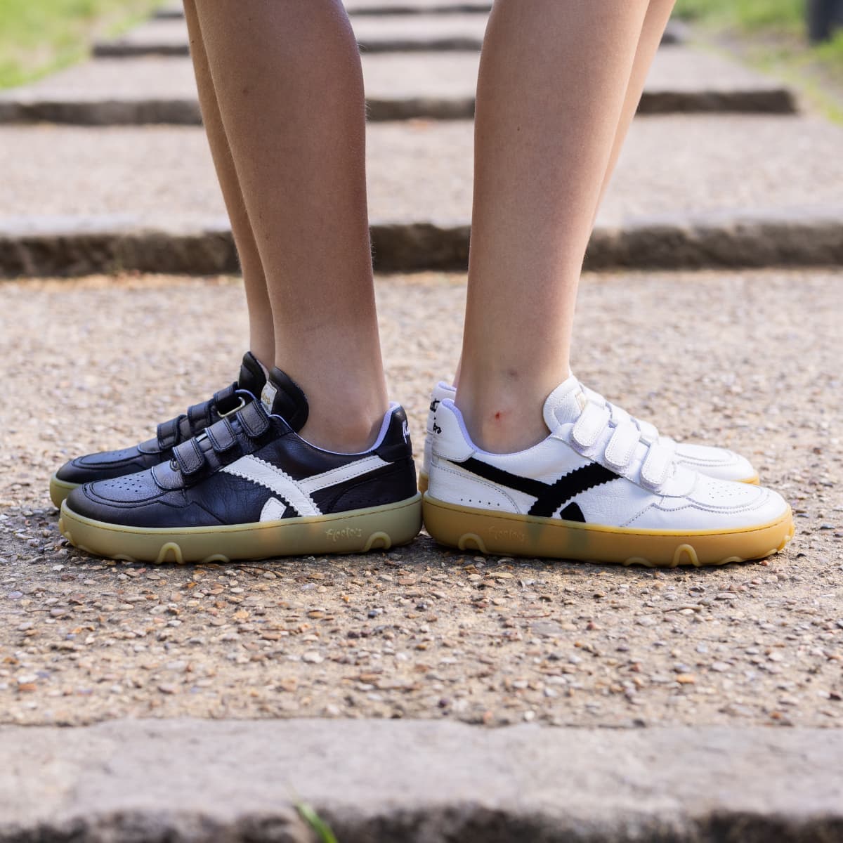 Two kids' low-top barefoot sneakers shown on feet standing on outdoor stone steps. One shoe is dark navy with white side stripes and a gum sole, and the other is white with black side stripes and a gum sole.