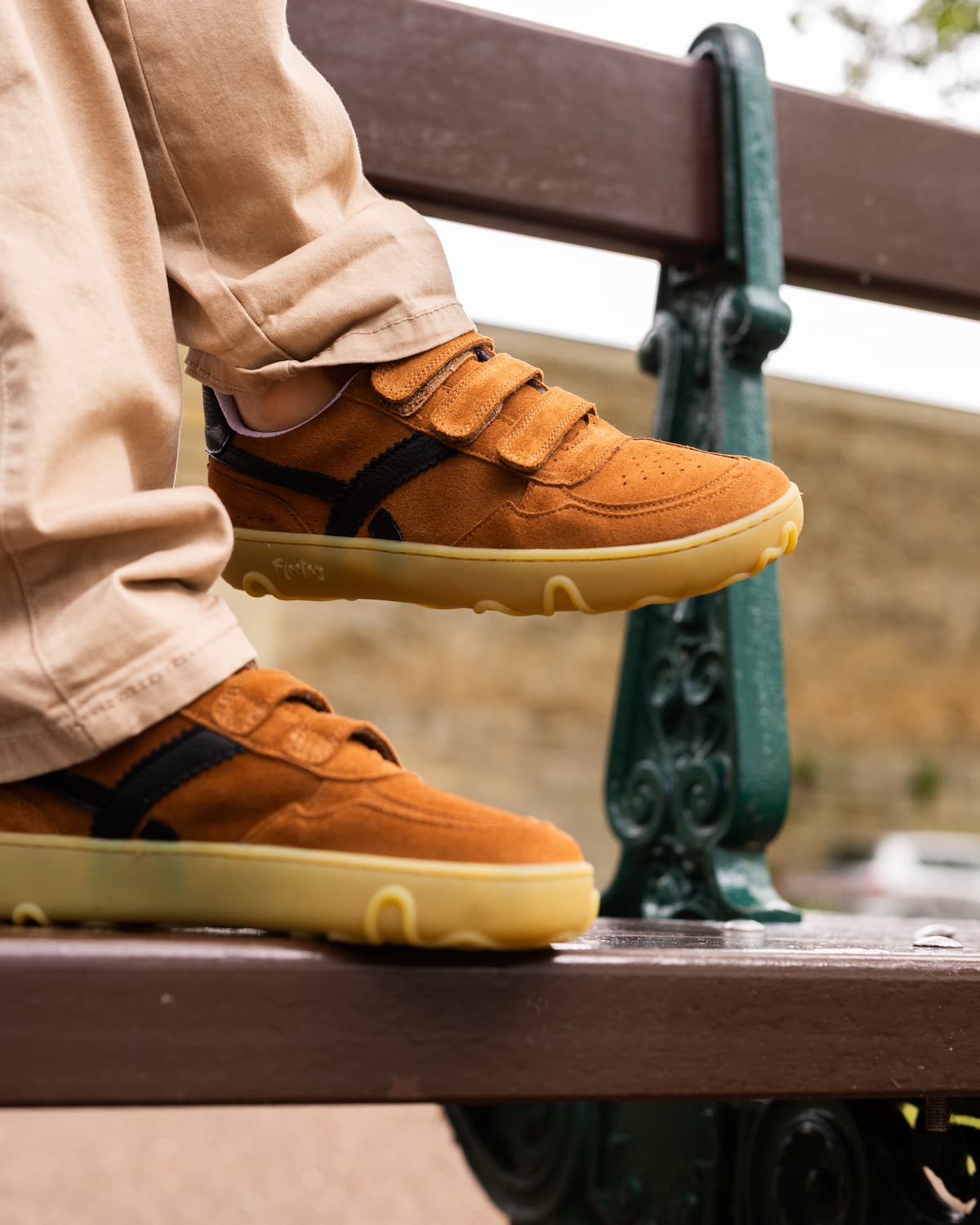 A kid sitting on a wooden bench wearing tan suede barefoot sneakers with double hook-and-loop straps, black side stripes, and gum soles, paired with beige pants.
