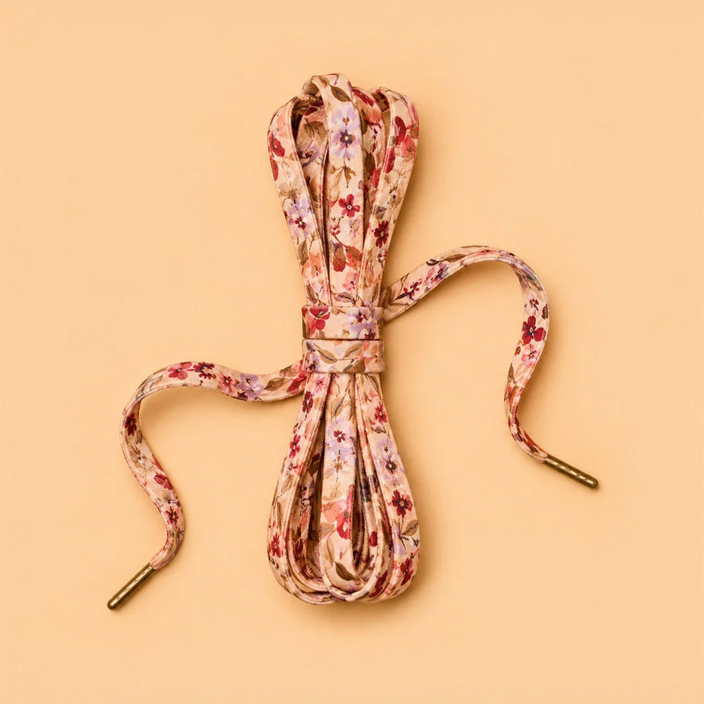 A coiled pair of floral lace shoelaces with gold metal tips, tied with a matching elastic band, on a solid peach background.