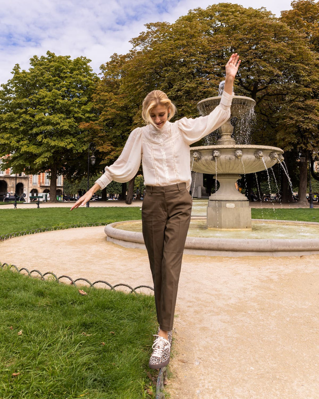 A woman posing outdoors in a park in front of a fountain, wearing a white long-sleeve blouse, brown trousers, and leopard print barefoot sneakers.