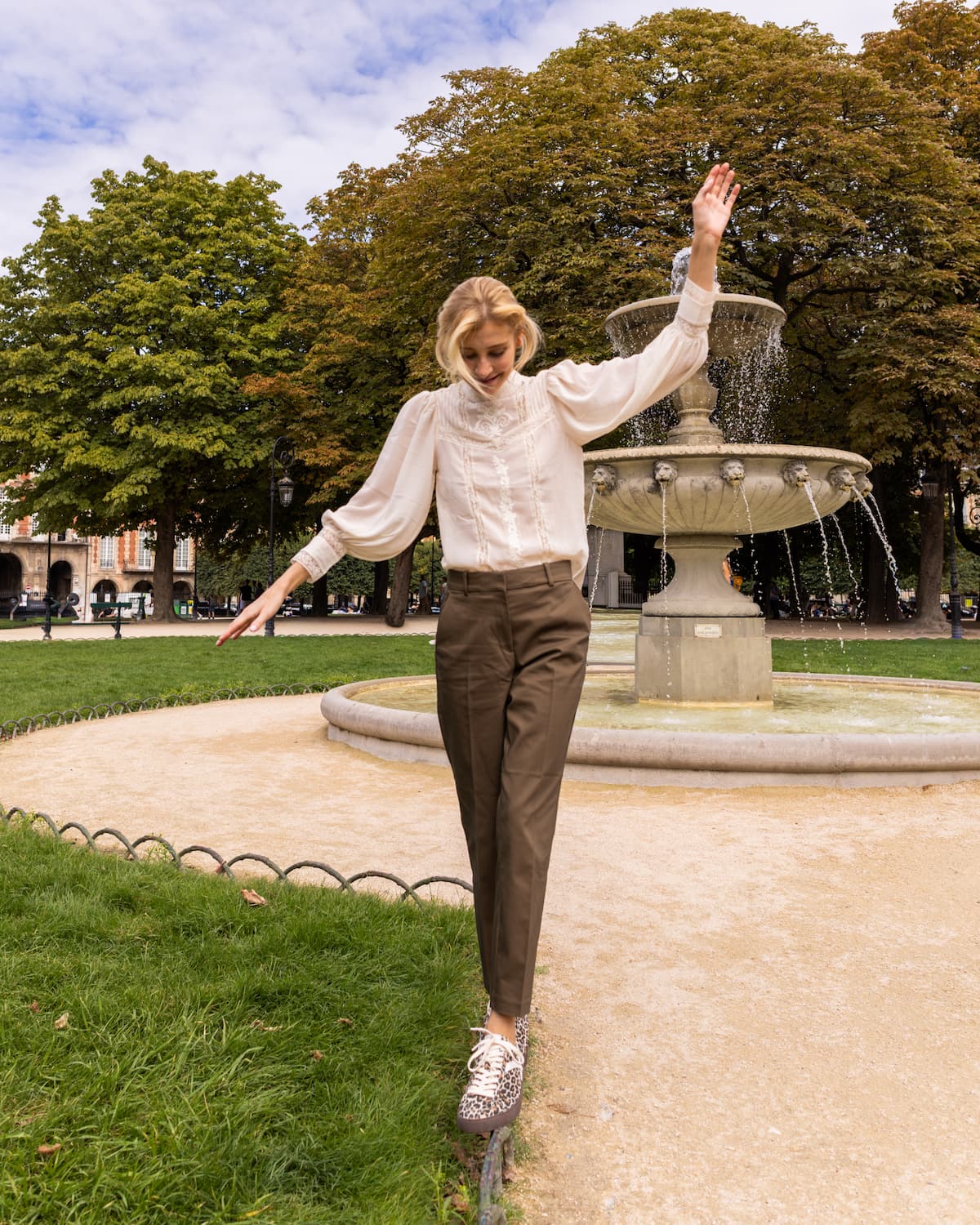 A woman posing outdoors in a park in front of a fountain, wearing a white long-sleeve blouse, brown trousers, and leopard print barefoot sneakers.