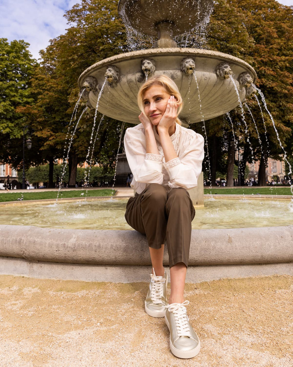 A woman sits on the edge of a fountain in an outdoor park, wearing a white long-sleeve blouse, brown cropped pants, and gold barefoot sneakers.