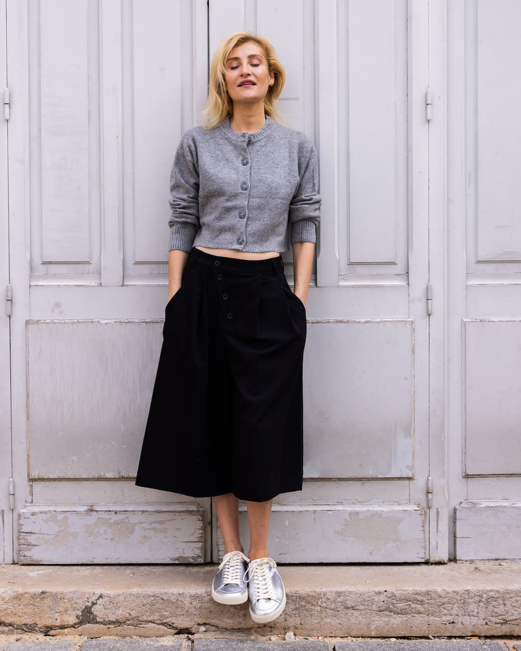 A woman stands in front of large weathered gray doors wearing a gray cropped button-front cardigan, a long black skirt, and silver barefoot sneakers.