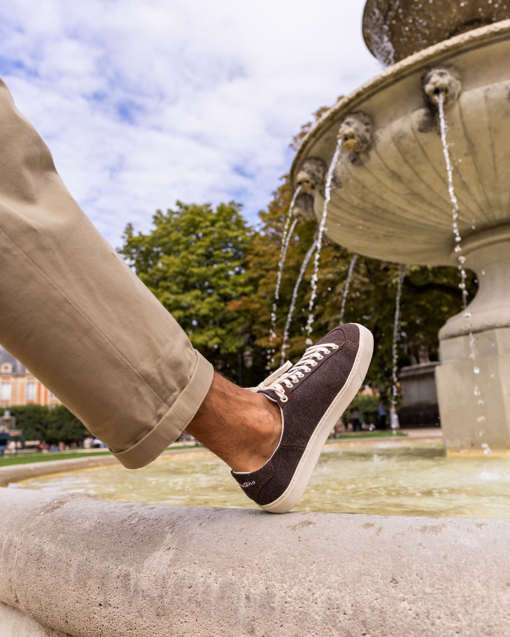 A person wearing a dark brown wool barefoot sneaker with white laces and a beige pant leg, standing on the edge of a stone fountain with water flowing in the background.