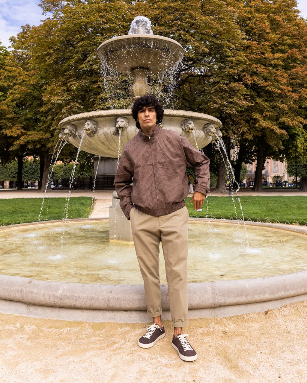 A man wearing a brown bomber jacket, beige trousers, and wool barefoot sneakers stands in front of a large stone fountain in a park with autumn trees.