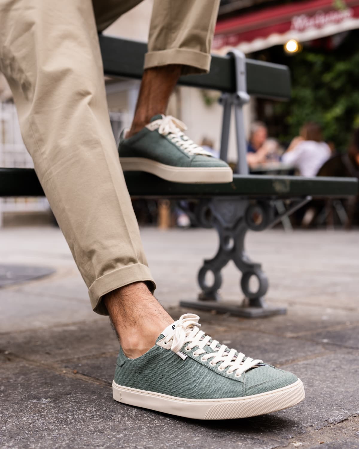 A person wearing sage green wool barefoot sneakers with off-white laces and soles, paired with beige trousers, sitting on a dark green bench in an outdoor cafe setting.